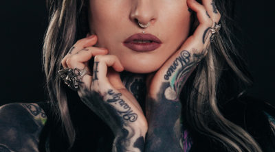 Woman with blonde hair & nose ring with tattoos hands & arms holding face - summer tattoo